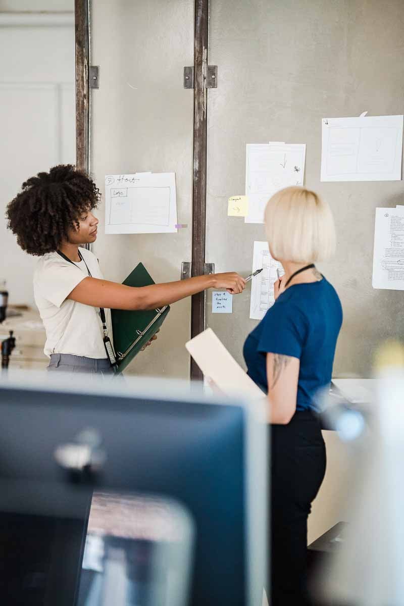 Two women in casual business attire stand in front of a wall in an office, pointing to papers taped to the wall.