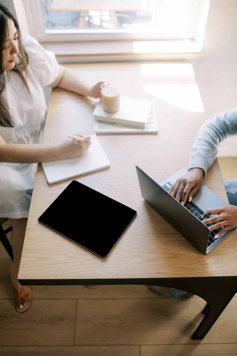 Two women seated a table working on laptop and writing in notebook with coffee and books stacked on table