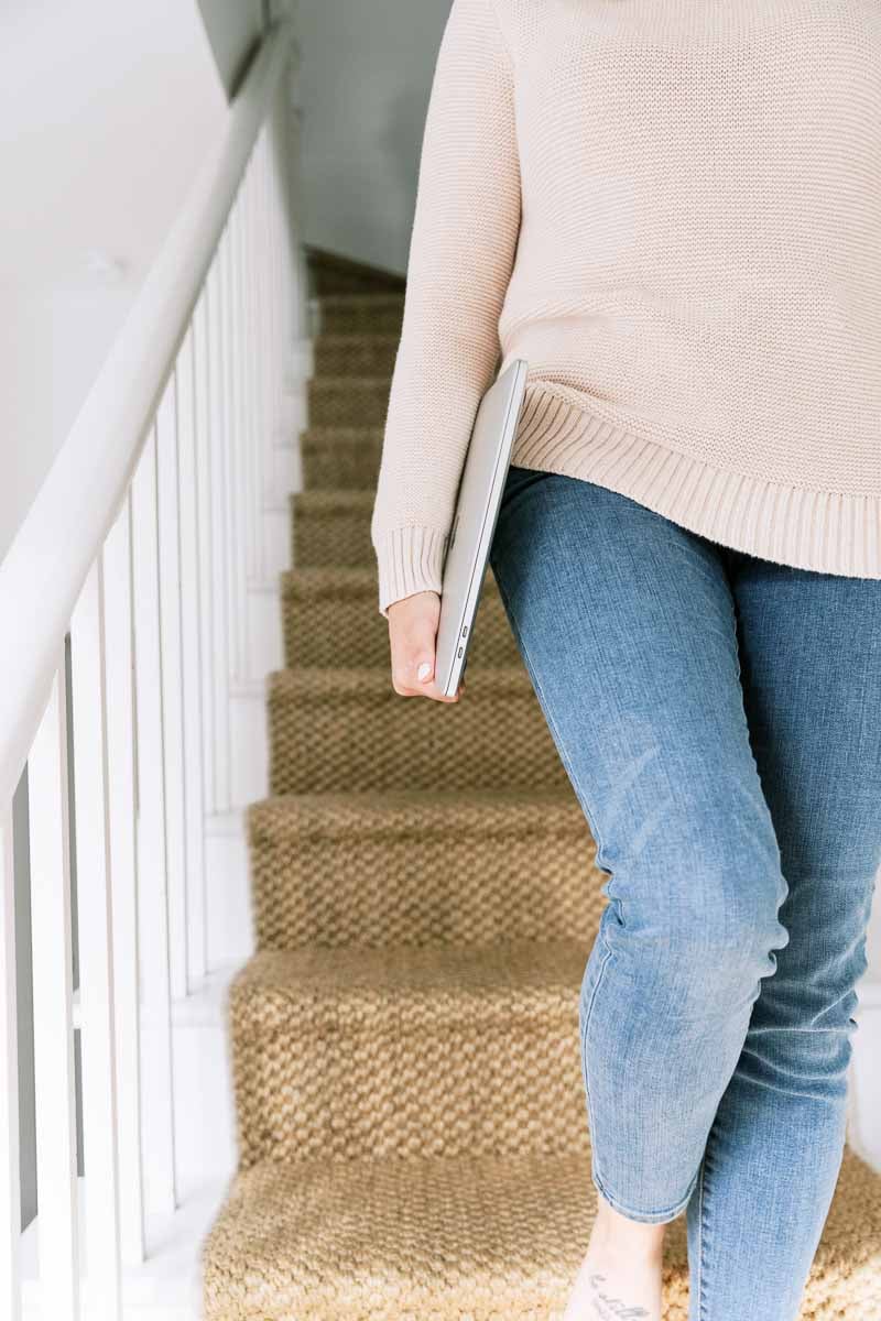 Elevae 23 006689 - Woman wearing a sweater and jeans holding a laptop and walking down carpeted stairs