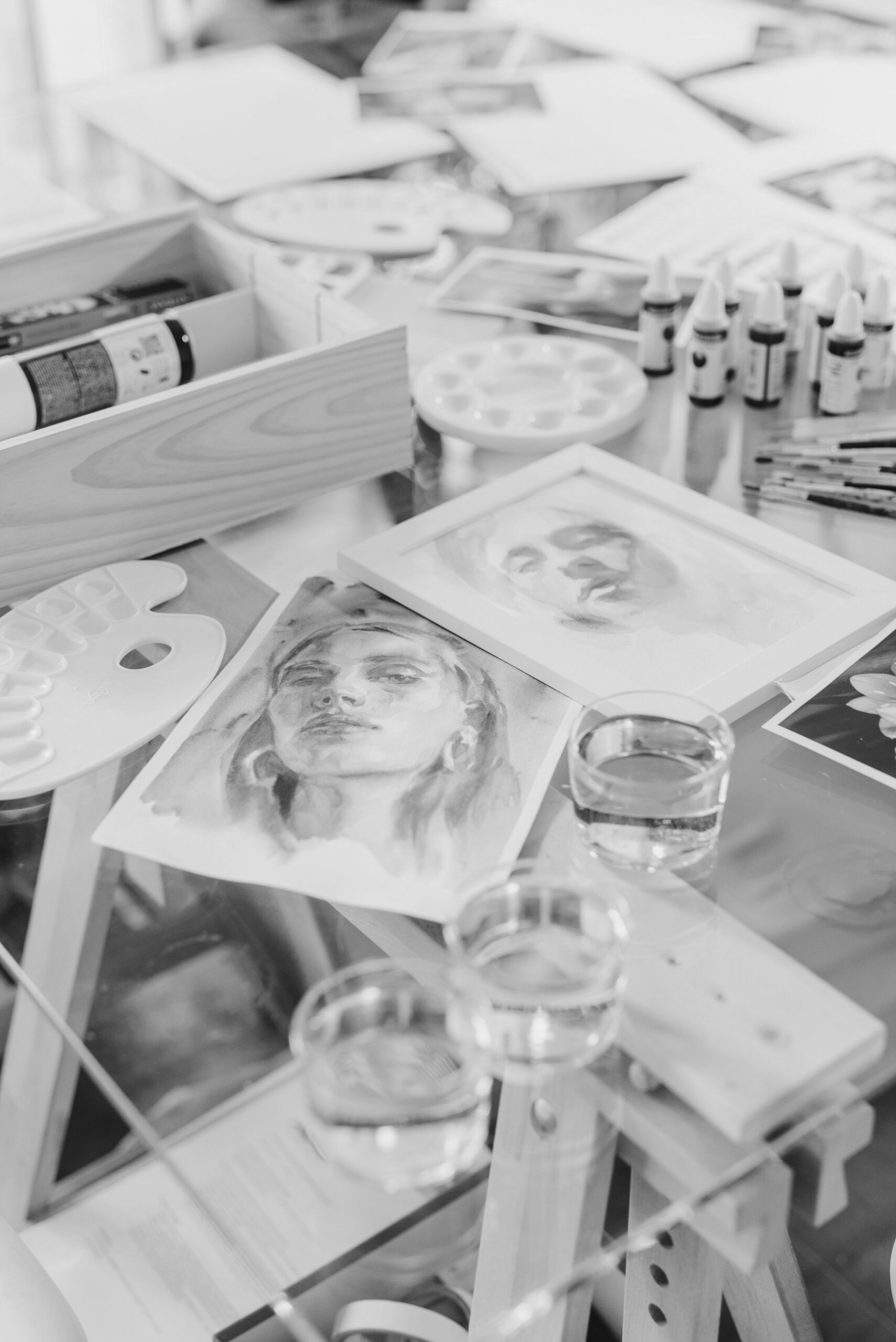 Art studio table with portrait sketches, paint palettes, brushes, ink bottles, and water glasses scattered across easels