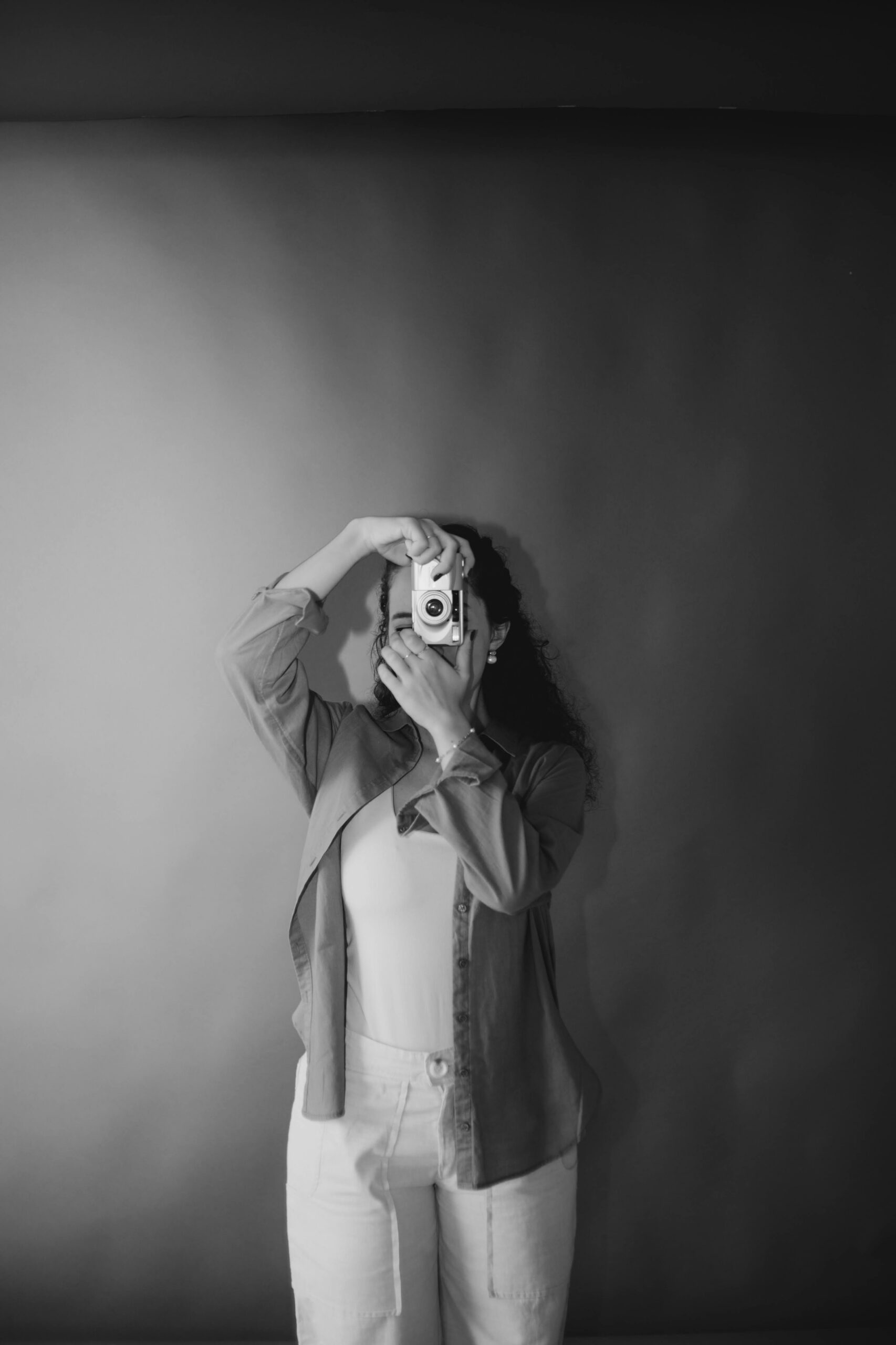 Woman holding a compact camera up to her face, obscuring it, in a black-and-white studio portrait