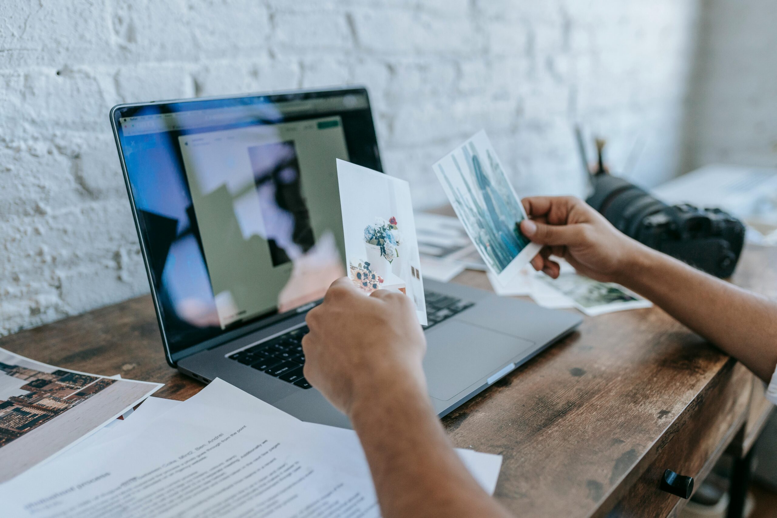 Photographer at a desk comparing printed photographs beside an open laptop and camera