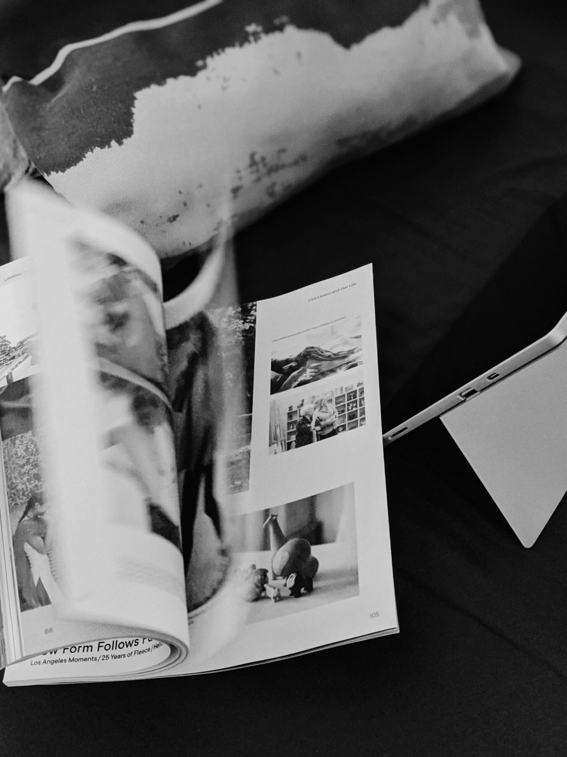 Black-and-white editorial photograph of an open design magazine beside a tablet on dark bedding